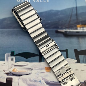 May include: A silver-toned watch band with a rectangular watch face. The band is made of multiple rectangular links. The image is set against a backdrop of a seascape and a table setting.