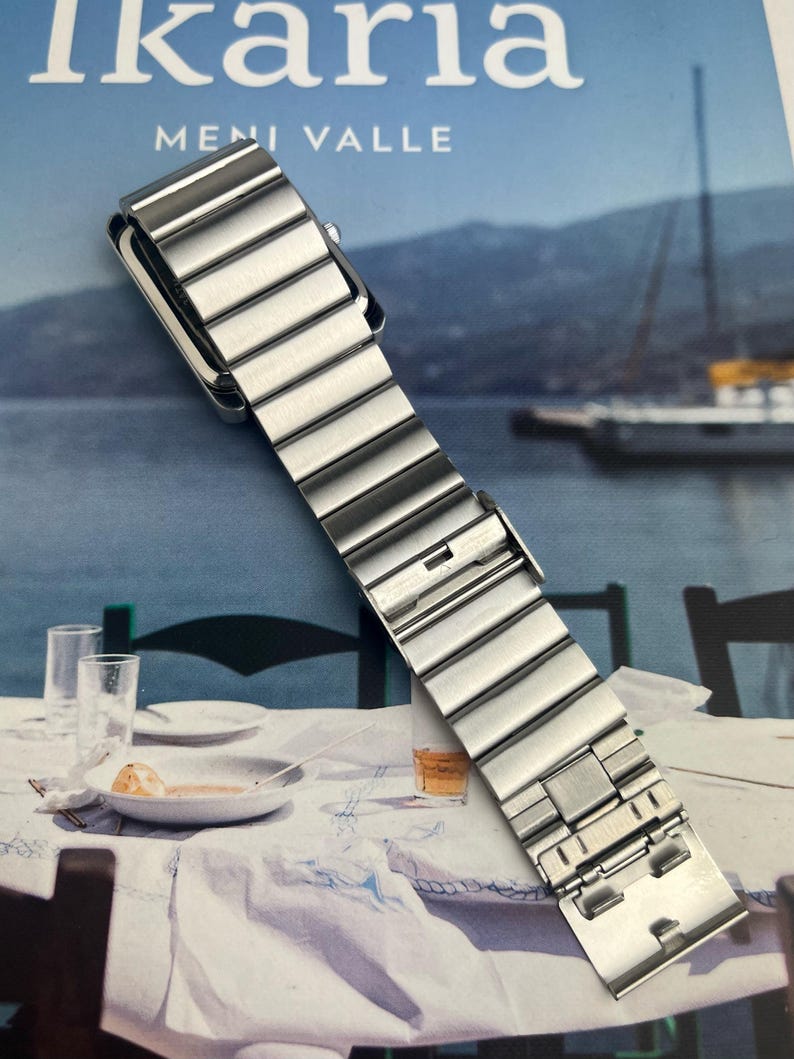 May include: A silver-toned watch band with a rectangular watch face. The band is made of multiple rectangular links. The image is set against a backdrop of a seascape and a table setting.