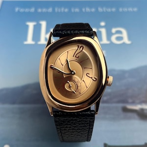May include: A gold-toned wristwatch with a black leather band. The watch face has a unique design with gold hands and a small seconds dial. The watch is set against a blurred background.
