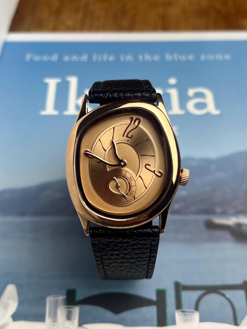 May include: A gold-toned wristwatch with a black leather band. The watch face has a unique design with gold hands and a small seconds dial. The watch is set against a blurred background.