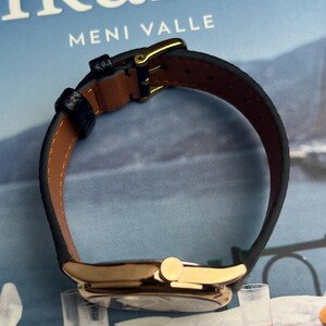 May include: A gold-toned wristwatch with a black leather band and a brown interior. The watch features a gold buckle and a round face. The text "Ikaria Meni Valle" is visible in the background.
