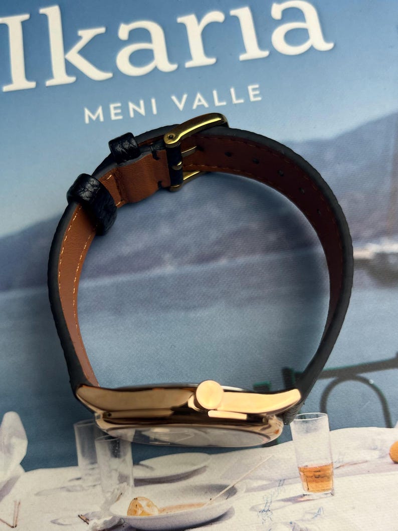 May include: A gold-toned wristwatch with a black leather band and a brown interior. The watch features a gold buckle and a round face. The text "Ikaria Meni Valle" is visible in the background.