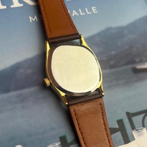 May include: A gold-toned wristwatch with a brown leather band. The watch has a rectangular face and a brown leather strap. The watch is displayed against a blurred background.
