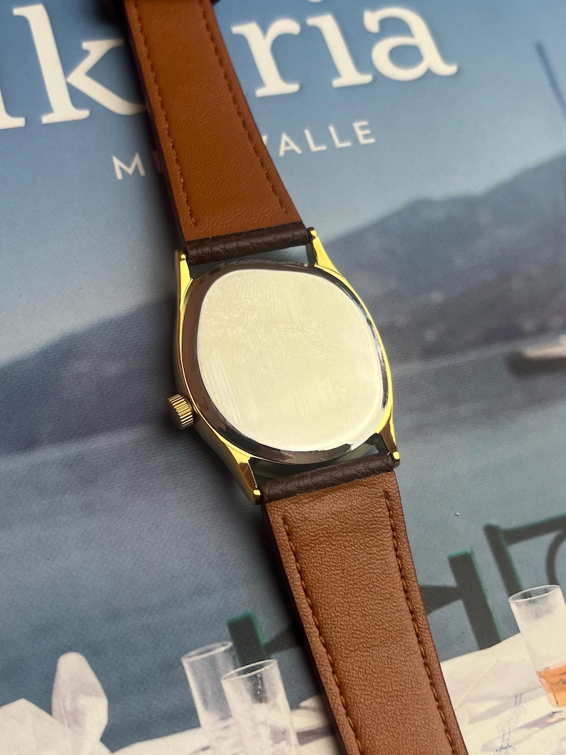 May include: A gold-toned wristwatch with a brown leather band. The watch has a rectangular face and a brown leather strap. The watch is displayed against a blurred background.