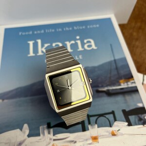 May include: A silver-toned wristwatch with a square black face and gold-colored trim. The watch has a silver band and displays the time. The watch is on a magazine with the word "Ikaria" on the cover.