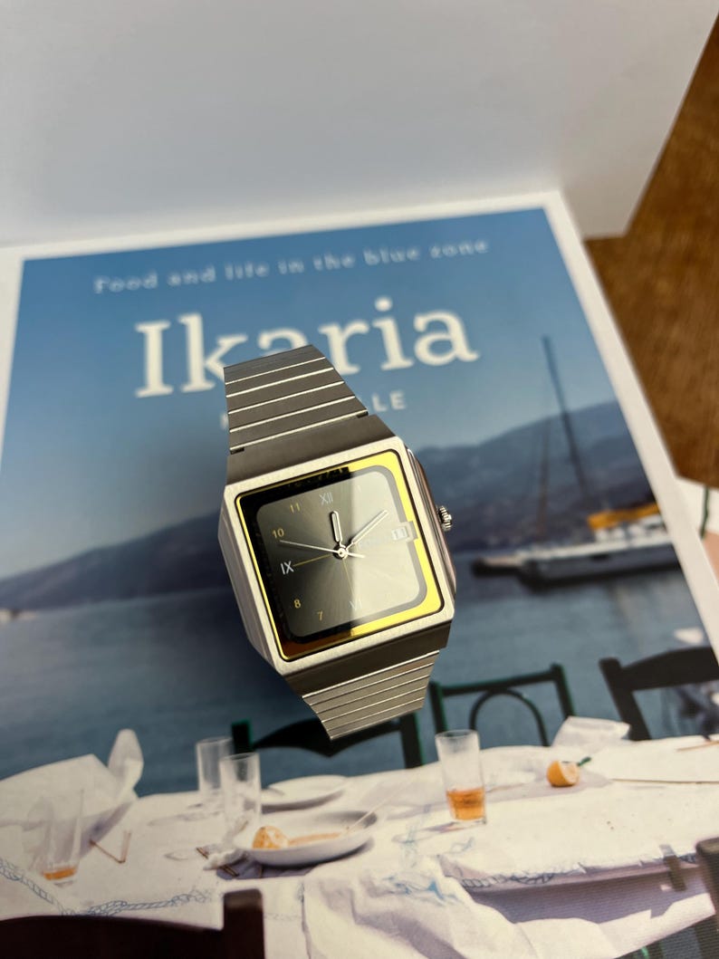 May include: A silver-toned wristwatch with a square black face and gold-colored trim. The watch has a silver band and displays the time. The watch is on a magazine with the word "Ikaria" on the cover.