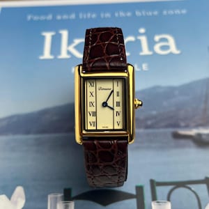 May include: A gold rectangular wristwatch with a brown leather strap. The watch face is cream-coloured with black Roman numerals and blue hands. The watch is labelled "Dimeina" and "Swiss".