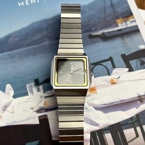 May include: A silver-toned wristwatch with a rectangular face and a matching metal band. The watch face has a gray background with black numerals and hands, and a yellow border. The watch is displayed on a magazine.