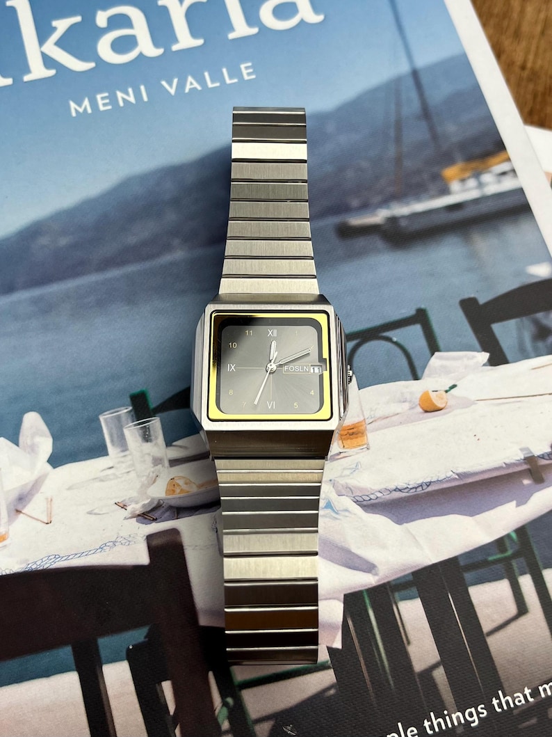 May include: A silver-toned wristwatch with a rectangular face and a matching metal band. The watch face has a gray background with black numerals and hands, and a yellow border. The watch is displayed on a magazine.