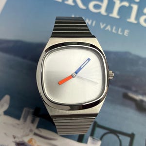 Puede incluir: Un reloj de pulsera plateado con esfera blanca y caja rectangular. El reloj tiene una manecilla azul y naranja, y una correa negra y plateada. El reloj está sobre un fondo borroso con la palabra "Ikaria".