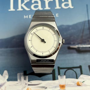 Peut inclure: Une montre-bracelet argent&eacute;e avec un cadran blanc et des aiguilles noires. La montre a un bo&icirc;tier rectangulaire et un bracelet en m&eacute;tal. Le texte "FOSLN QUARTA WATCH" est visible sur le cadran. En arri&egrave;re-plan, un magazine avec le mot "Ikaria".
