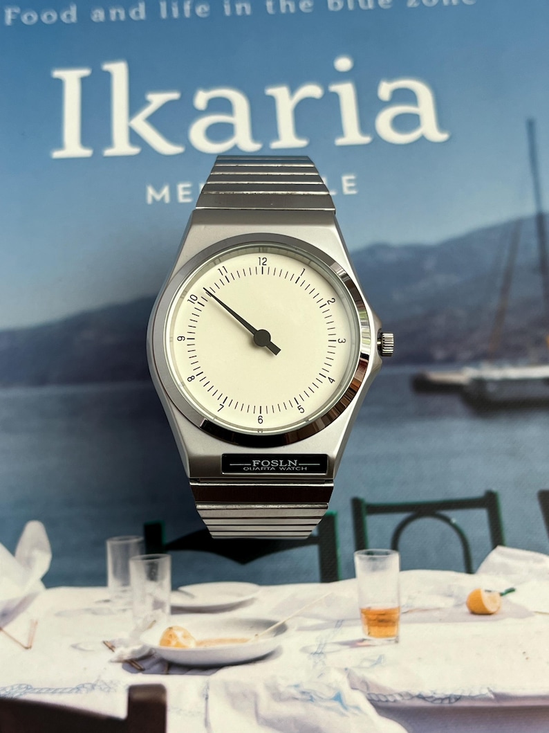 Peut inclure: Une montre-bracelet argent&eacute;e avec un cadran blanc et des aiguilles noires. La montre a un bo&icirc;tier rectangulaire et un bracelet en m&eacute;tal. Le texte "FOSLN QUARTA WATCH" est visible sur le cadran. En arri&egrave;re-plan, un magazine avec le mot "Ikaria".