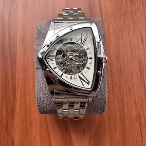 May include: A silver-toned triangular wristwatch with a skeleton dial revealing internal mechanical gears. The watch features a metallic link bracelet and a white face with black Roman numeral hour markers at the three, six, and twelve positions.
