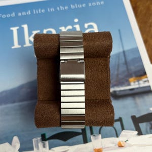 May include: A silver-toned watch band with a brushed metal finish is displayed on a brown, textured surface. The band is composed of rectangular links and is partially closed. The background features text that reads "Food and life in the blue zone" and the word "Ikaria".