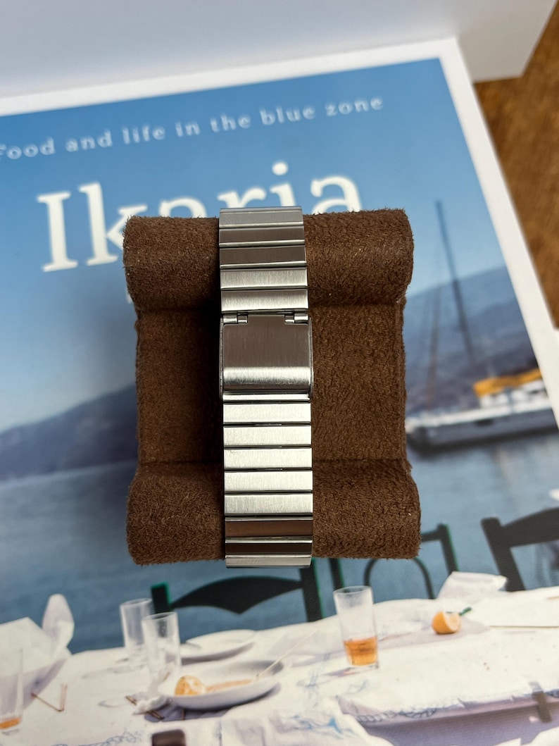 May include: A silver-toned watch band with a brushed metal finish is displayed on a brown, textured surface. The band is composed of rectangular links and is partially closed. The background features text that reads "Food and life in the blue zone" and the word "Ikaria".