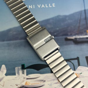 May include: A silver-toned digital watch with a rectangular face and a segmented metal band. The watch face displays a digital time readout. The band is composed of rectangular links.