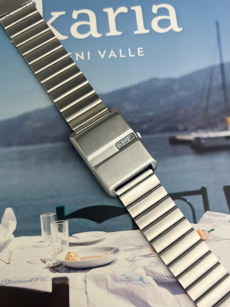 May include: A silver-toned digital watch with a rectangular face and a segmented metal band. The watch face displays a digital time readout. The band is composed of rectangular links.