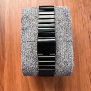Peut inclure: Un bracelet de montre en m&eacute;tal noir mat avec un fermoir pliant rectangulaire, enroul&eacute; autour d'un coussin de pr&eacute;sentation en tissu gris textur&eacute;. Le bracelet se compose de maillons rectangulaires horizontaux reli&eacute;s en cha&icirc;ne flexible.