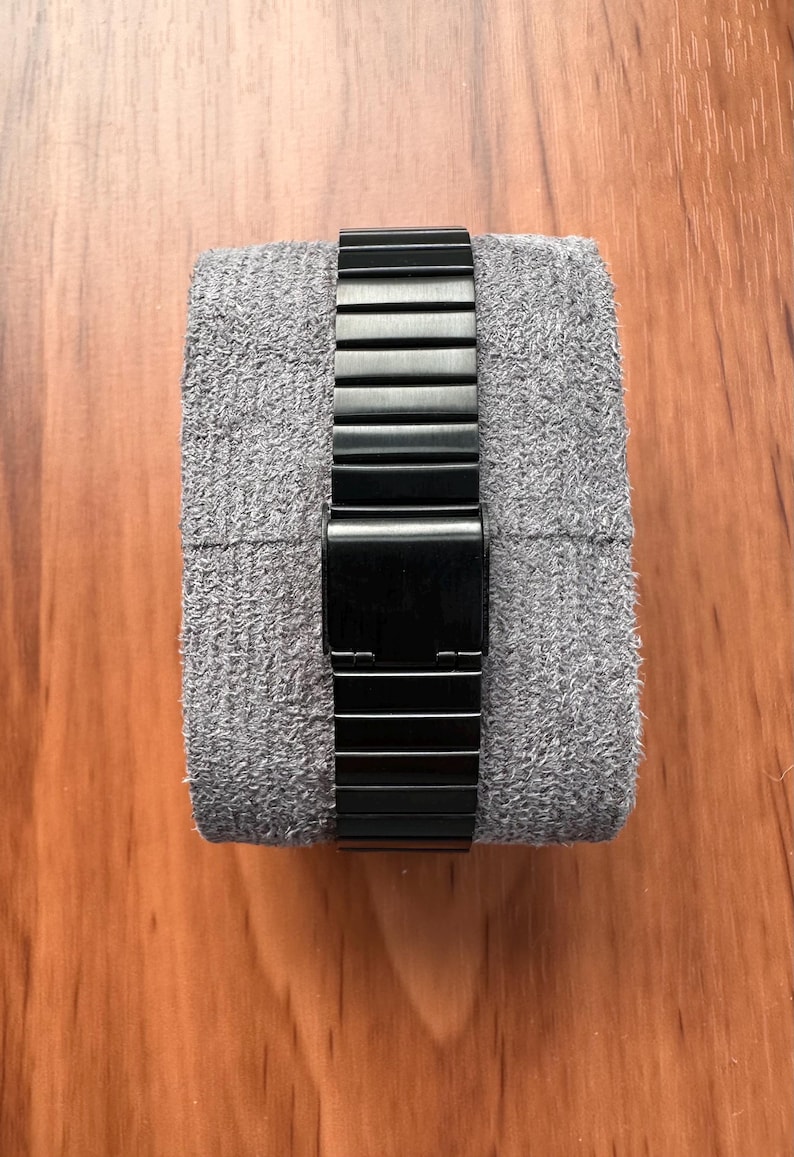 Peut inclure: Un bracelet de montre en m&eacute;tal noir mat avec un fermoir pliant rectangulaire, enroul&eacute; autour d'un coussin de pr&eacute;sentation en tissu gris textur&eacute;. Le bracelet se compose de maillons rectangulaires horizontaux reli&eacute;s en cha&icirc;ne flexible.