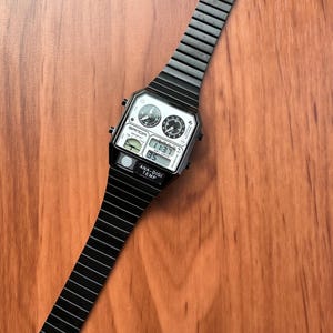 Peut inclure: Une montre-bracelet analogique et num&eacute;rique noire avec un cadran rectangulaire, deux petits cadrans analogiques circulaires, un affichage num&eacute;rique indiquant 11:37, et le texte SANDA et ANA-DIGI TEMP. Elle poss&egrave;de un bracelet &agrave; maillons noir.