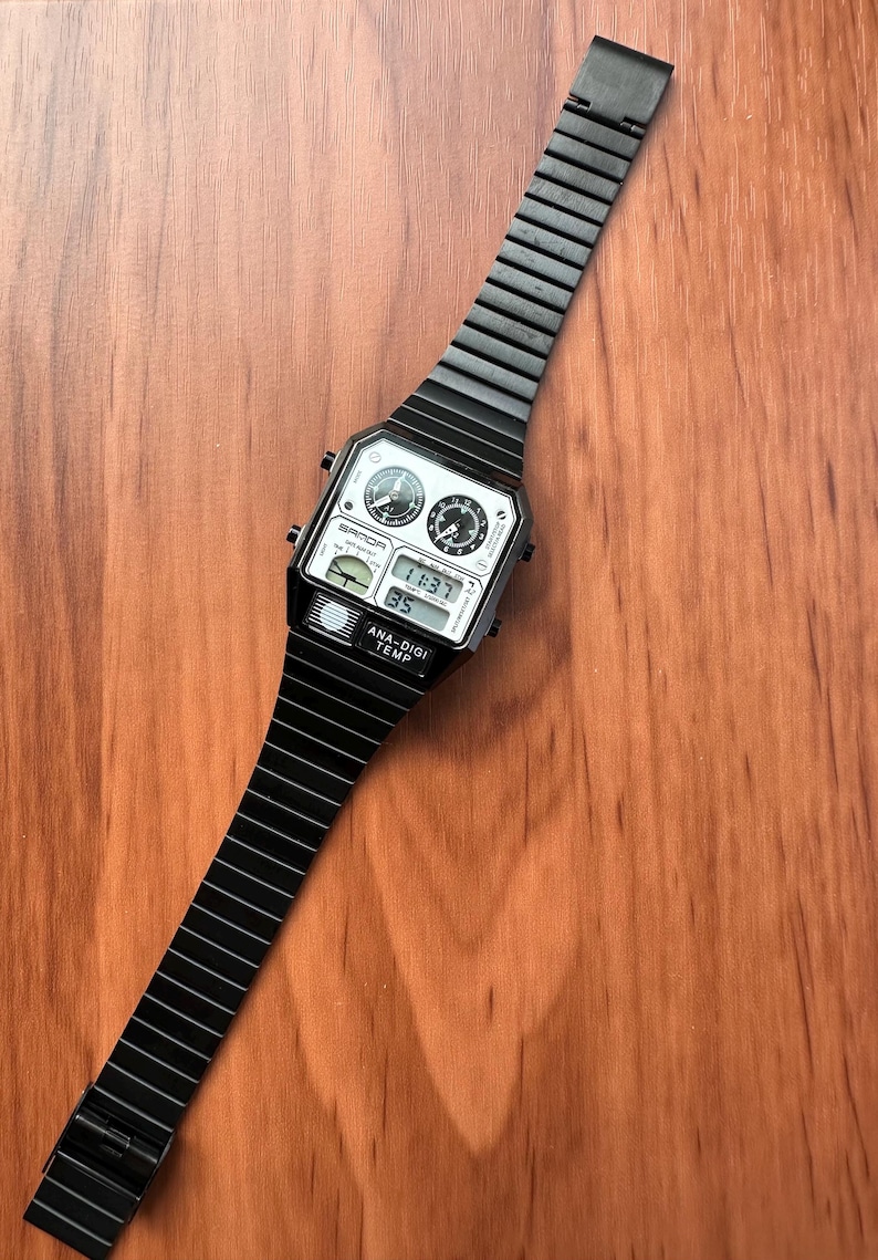 Peut inclure: Une montre-bracelet analogique et num&eacute;rique noire avec un cadran rectangulaire, deux petits cadrans analogiques circulaires, un affichage num&eacute;rique indiquant 11:37, et le texte SANDA et ANA-DIGI TEMP. Elle poss&egrave;de un bracelet &agrave; maillons noir.