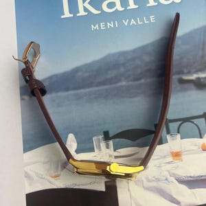May include: A gold-toned wristwatch with a brown leather band is displayed on a book titled "Ikaria". The watch has a rectangular face and a classic design. The book cover features the title in white letters against a blue background.