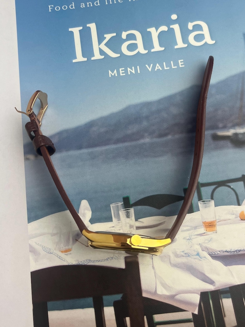 May include: A gold-toned wristwatch with a brown leather band is displayed on a book titled "Ikaria". The watch has a rectangular face and a classic design. The book cover features the title in white letters against a blue background.