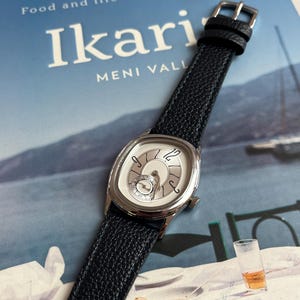 May include: A silver-toned wristwatch with a white face and black leather band. The watch has a unique oval shape and a textured black band. The watch is resting on a book titled "Ikari" with the text "Food and life in the blue zone".
