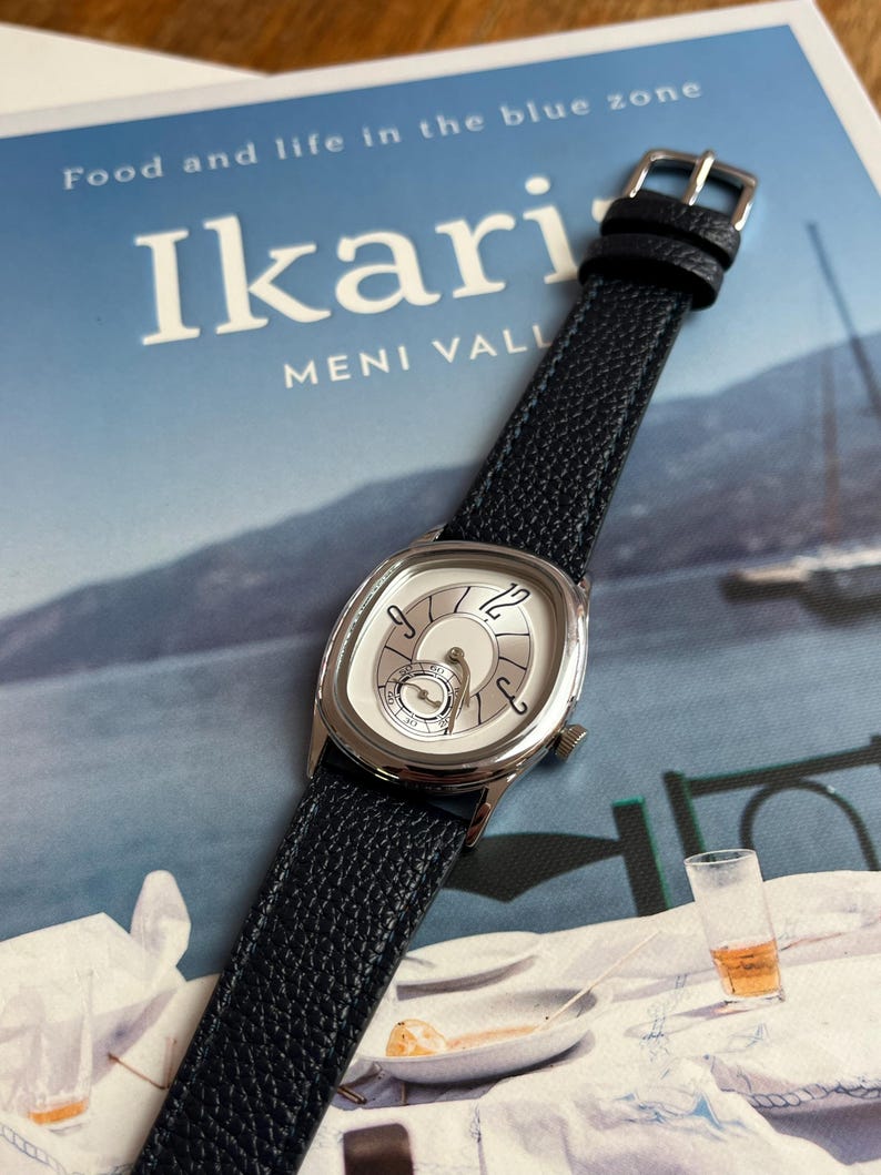 May include: A silver-toned wristwatch with a white face and black leather band. The watch has a unique oval shape and a textured black band. The watch is resting on a book titled "Ikari" with the text "Food and life in the blue zone".