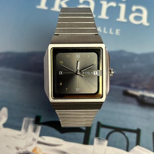 May include: A silver-toned wristwatch with a square face and a matching band. The watch face displays Roman numerals and the text "FOSLN 11". The background features the word "Ikaria" and a blurred image.