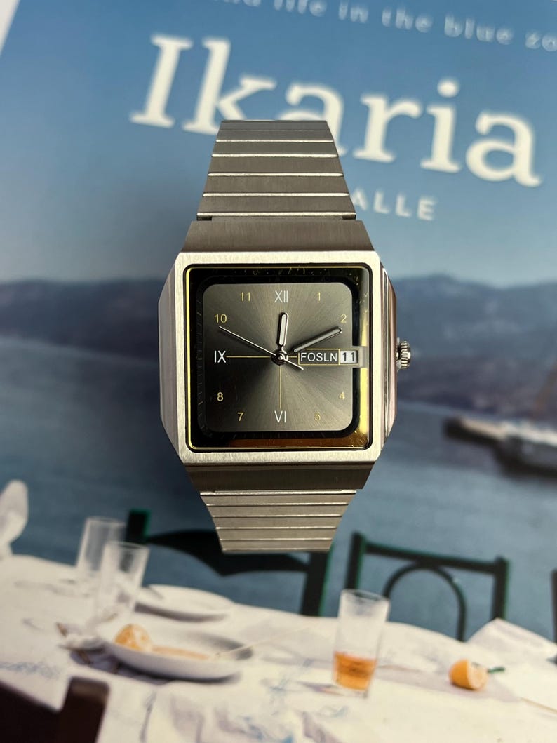 May include: A silver-toned wristwatch with a square face and a matching band. The watch face displays Roman numerals and the text "FOSLN 11". The background features the word "Ikaria" and a blurred image.