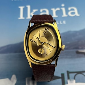 May include: A gold-toned wristwatch with a brown leather band. The watch face has a unique shape and features gold hands and a small seconds dial. The watch is set against a blurred background with the word "Ikaria" visible.