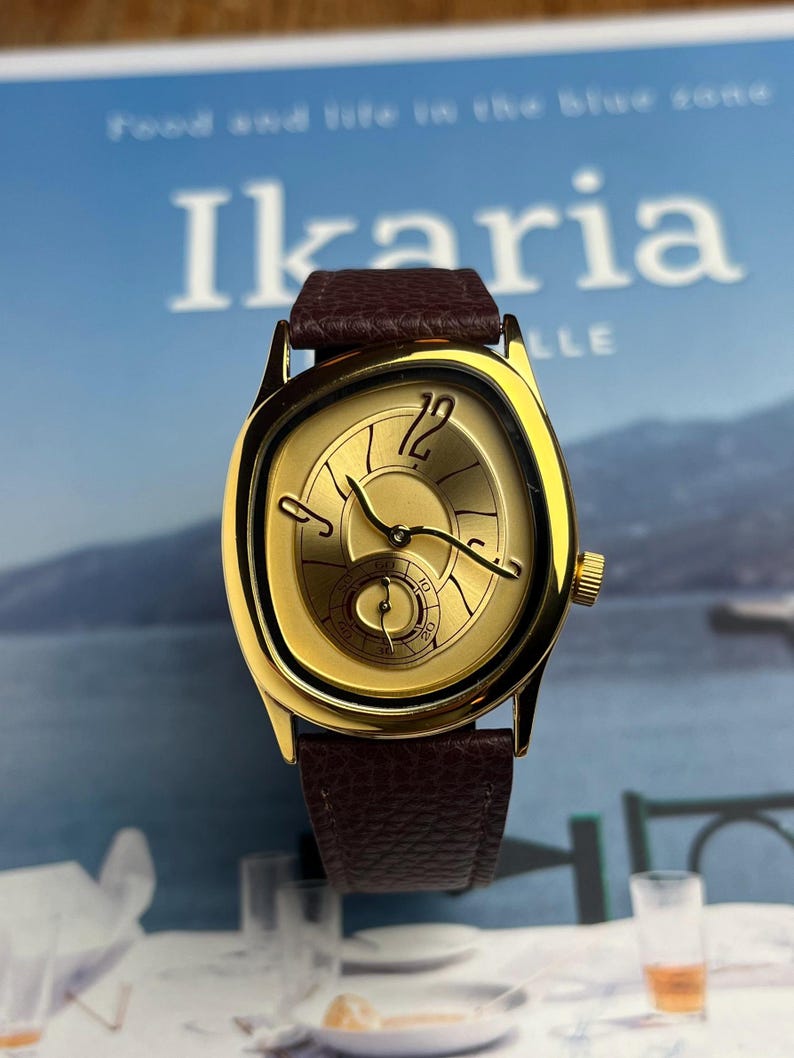 May include: A gold-toned wristwatch with a brown leather band. The watch face has a unique shape and features gold hands and a small seconds dial. The watch is set against a blurred background with the word "Ikaria" visible.