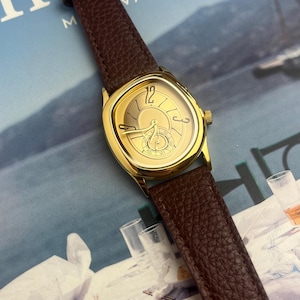 May include: A gold-toned wristwatch with a brown leather band. The watch face has a unique shape and features gold hands and markers. The band is a rich brown color, and the watch is set against a blurred background.