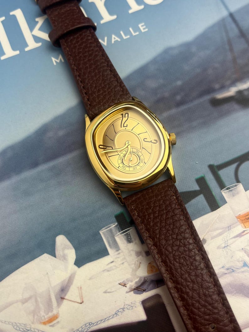 May include: A gold-toned wristwatch with a brown leather band. The watch face has a unique shape and features gold hands and markers. The band is a rich brown color, and the watch is set against a blurred background.
