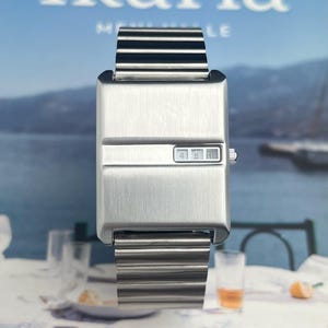 May include: A rectangular, brushed metal wristwatch with a digital display showing the time. The watch has a matching metal band. The word "Ikaria" is visible in the background. The watch is displayed on a table with glasses and a slice of orange.
