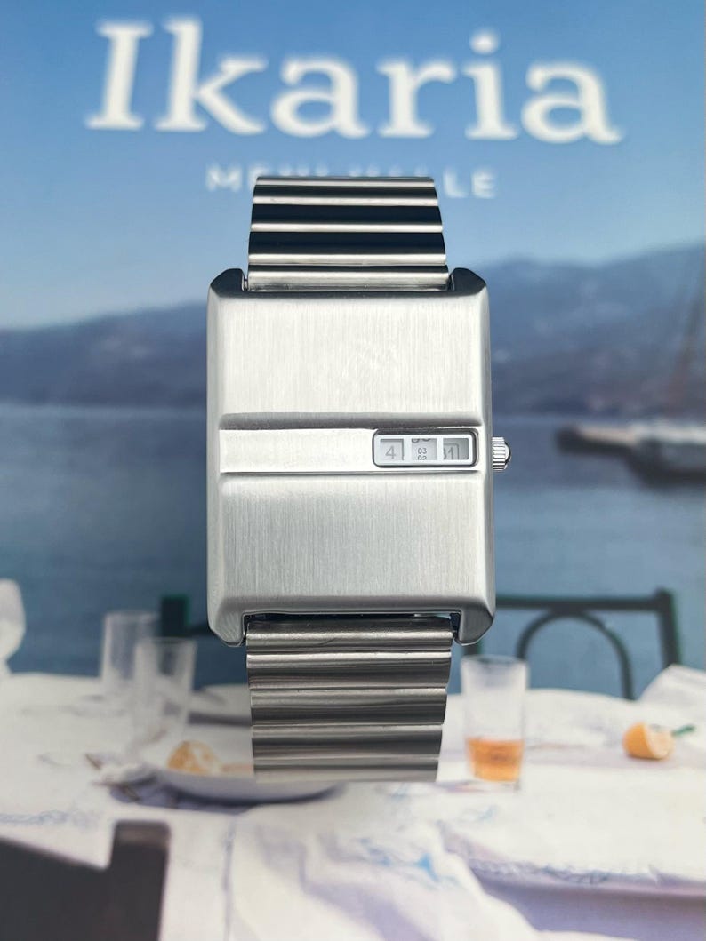 May include: A rectangular, brushed metal wristwatch with a digital display showing the time. The watch has a matching metal band. The word "Ikaria" is visible in the background. The watch is displayed on a table with glasses and a slice of orange.