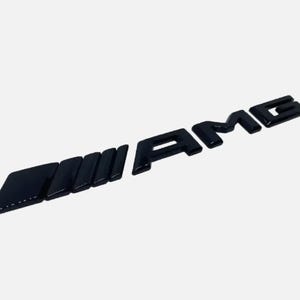 May include: Black AMG emblem with a glossy finish. The emblem features the letters "AMG" in a stylized font, with a series of angled rectangular shapes to the left of the letters. The emblem is designed for automotive use.