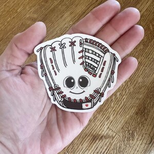 Może przedstawiać: Biała naklejka z rękawicą do baseballu z uśmiechniętą twarzą, dużymi czarnymi oczami i czerwonymi przeszyciami. Naklejka ma brązowy mankiet i mały czerwony detal w kształcie serca. Naklejka jest trzymana w dłoni.