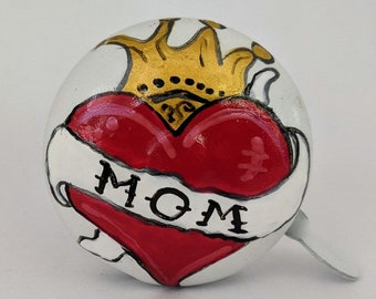 May include: White bicycle bell with a red heart and a gold crown. The heart has a banner that says "MOM".