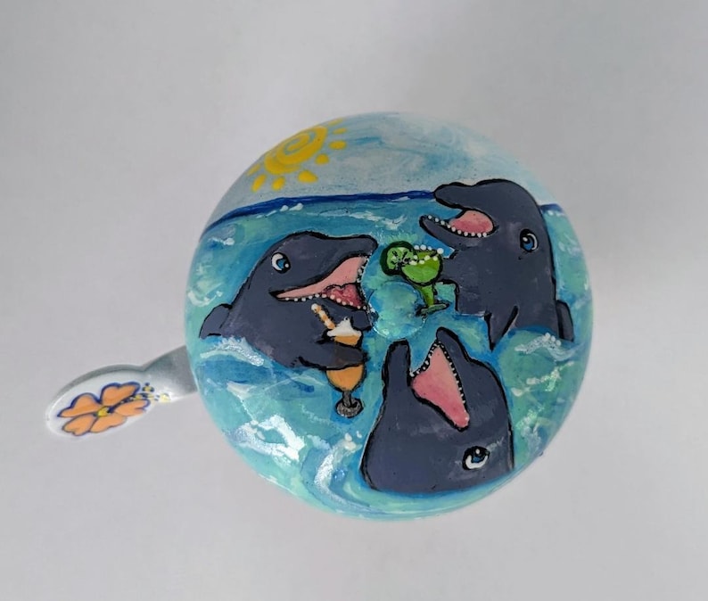 Puede incluir: Una campanilla de bicicleta de cer&aacute;mica pintada a mano con una escena oce&aacute;nica azul, verde y blanca. Se representan tres delfines nadando y sosteniendo bebidas. Un sol amarillo se encuentra en la esquina superior izquierda.