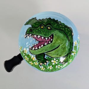 Op de afbeelding: Een ronde fietsbel met een lichtblauwe achtergrond voorzien van een handgeschilderde groene alligator met een open bek en witte tanden. De alligator is omringd door kleine witte bloemetjes met gele hartjes en groene steeltjes onderaan.