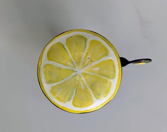 May include: A round bicycle bell featuring a vibrant yellow and white lemon slice graphic on its top surface. The design displays a detailed citrus pattern with distinct segments radiating from the center, paired with a black metal lever arm.