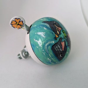 Puede incluir: Una campanilla de bicicleta blanca con una escena de oc&eacute;ano azul y verde pintada en ella. Dos delfines negros y grises nadan en el agua. Una flor amarilla con un centro negro est&aacute; en el lado de la campanilla.