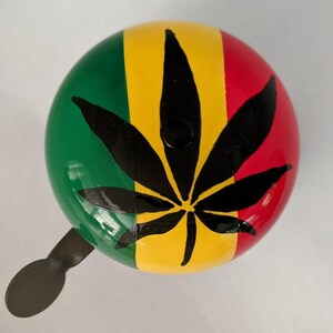 Puede incluir: Una campana de bicicleta verde, amarilla y roja con un diseño de hoja de marihuana negra.