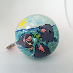 Puede incluir: Un adorno de cer&aacute;mica pintado a mano con tres delfines disfrutando de una bebida tropical en el oc&eacute;ano. El adorno es azul, verde y amarillo con un fondo blanco.