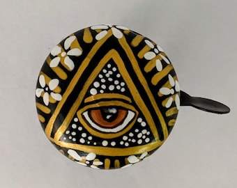May include: A black and gold bicycle bell with a white all-seeing eye design. The eye is surrounded by white flowers and a black and gold geometric pattern.