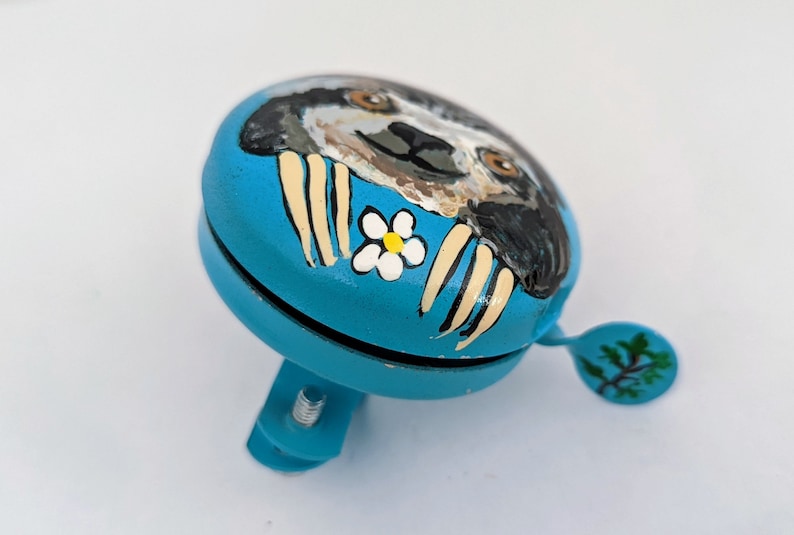 Op de afbeelding: Een blauwe fietsbel met een geschilderd luiaardontwerp. De luiaard heeft een witte bloem op zijn borst en is geschilderd in tinten bruin, zwart en wit.