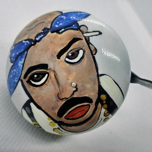 Puede incluir: Una campanilla de bicicleta blanca con un retrato pintado a mano de una persona que lleva un pañuelo azul. La persona tiene piel morena, ojos marrones y bigote. La campanilla tiene un anillo de metal negro.