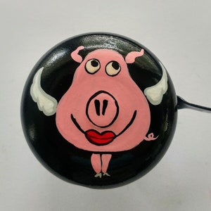 Op de afbeelding: Een zwarte fietsbel met een geschilderd roze varken met witte engelenvleugels en rode lippenstift. Het varken heeft een zwarte omtrek en zwarte ogen.